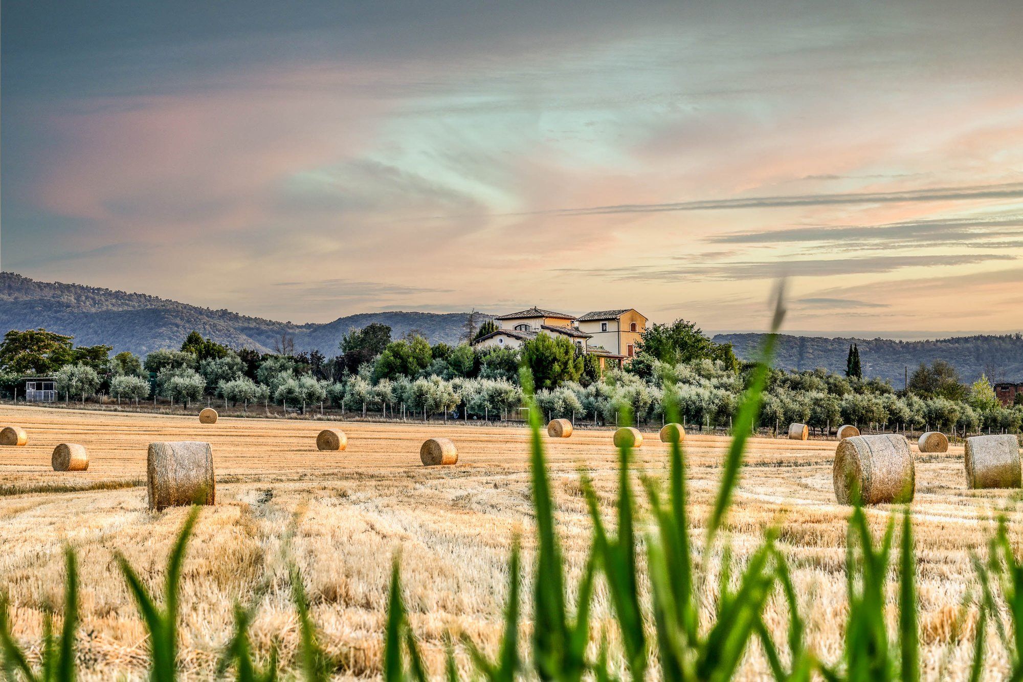 Il casale sulla collina al tramonto, dietro un campo di grano mietuto con le balle di fieno