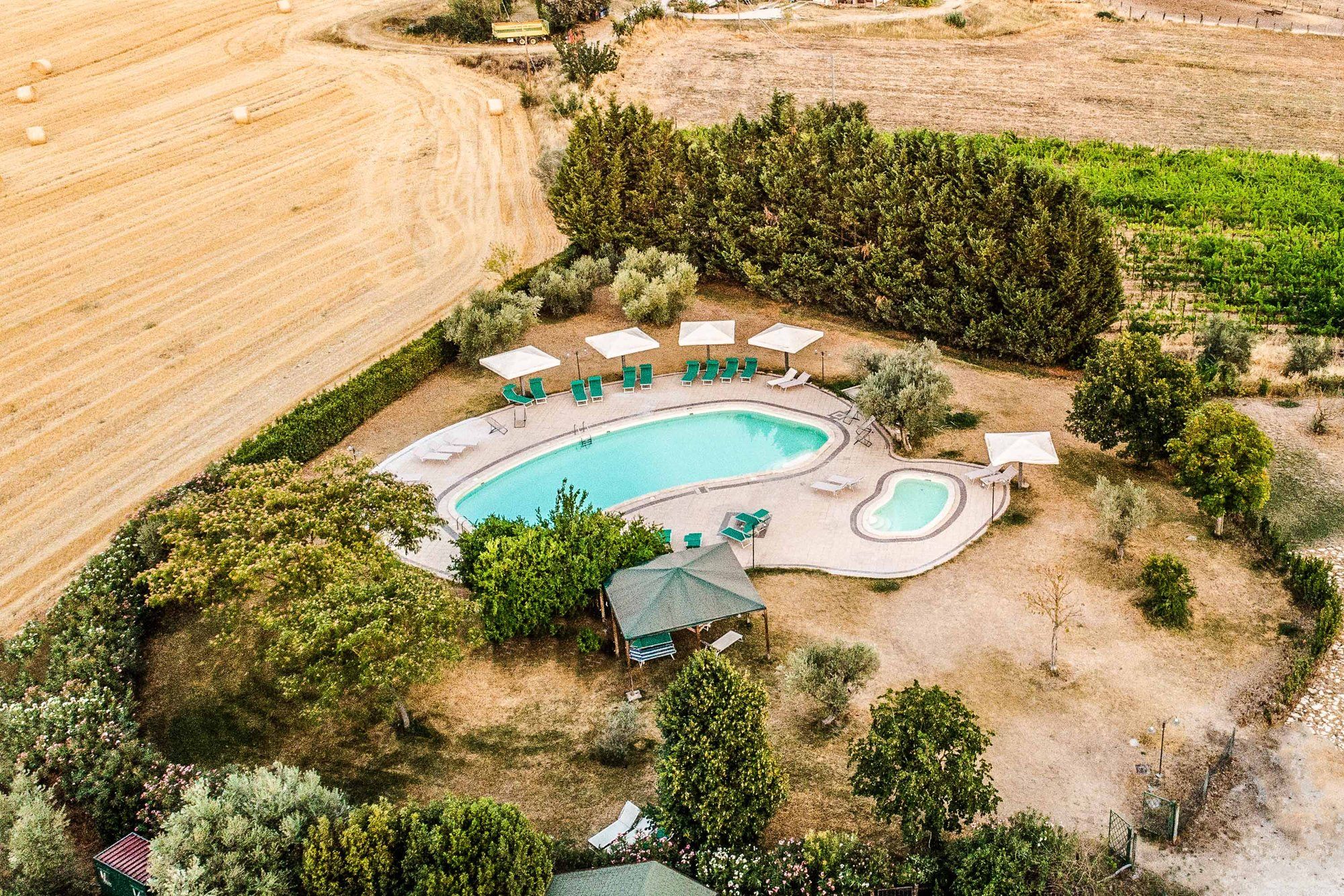 La piscina per adulti e la piscina per bambini viste dall'alto, con lettini e ombrelloni, tra gli ulivi e i campi